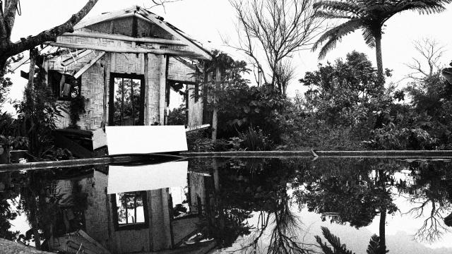 an black and white image of a bamboo hut with reflection over water in a pool. 